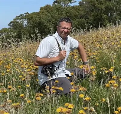 Mauricio Bonifacino, profesor de botánica, sentado en un campo florido con plantas nativas, sosteniendo una cámara.
